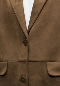 Blazer en daim marron avec deux boutons et poches à rabat, porté sur une chemise blanche.