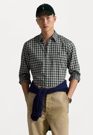 Male wearing green plaid button-up shirt with sleeves rolled, navy sweater tied around waist, beige pants, green cap, and wristwatch, hands in pockets.