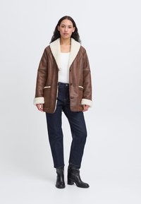 Giubbotto in pelle marrone con colletto in shearling, caratterizzato da una vestibilità rilassata, due tasche e cuciture bianche a contrasto. Abbinato a jeans scuri e stivali.