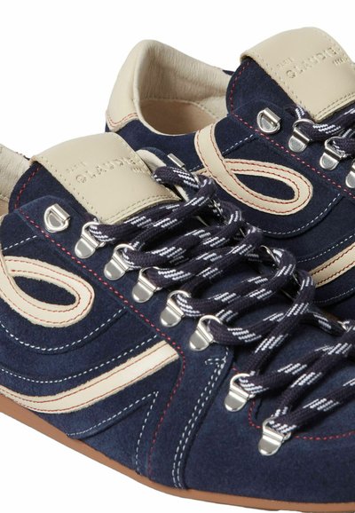 Baskets en daim bleu marine avec des accents en cuir beige, des coutures rouges et blanches, des lacets à motifs et des œillets en métal, montrant un détail en gros plan.