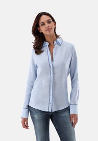 Light blue linen shirt with long sleeves, button-up front, contrasting collar and cuff trim, and a relaxed fit. Paired with distressed denim jeans.