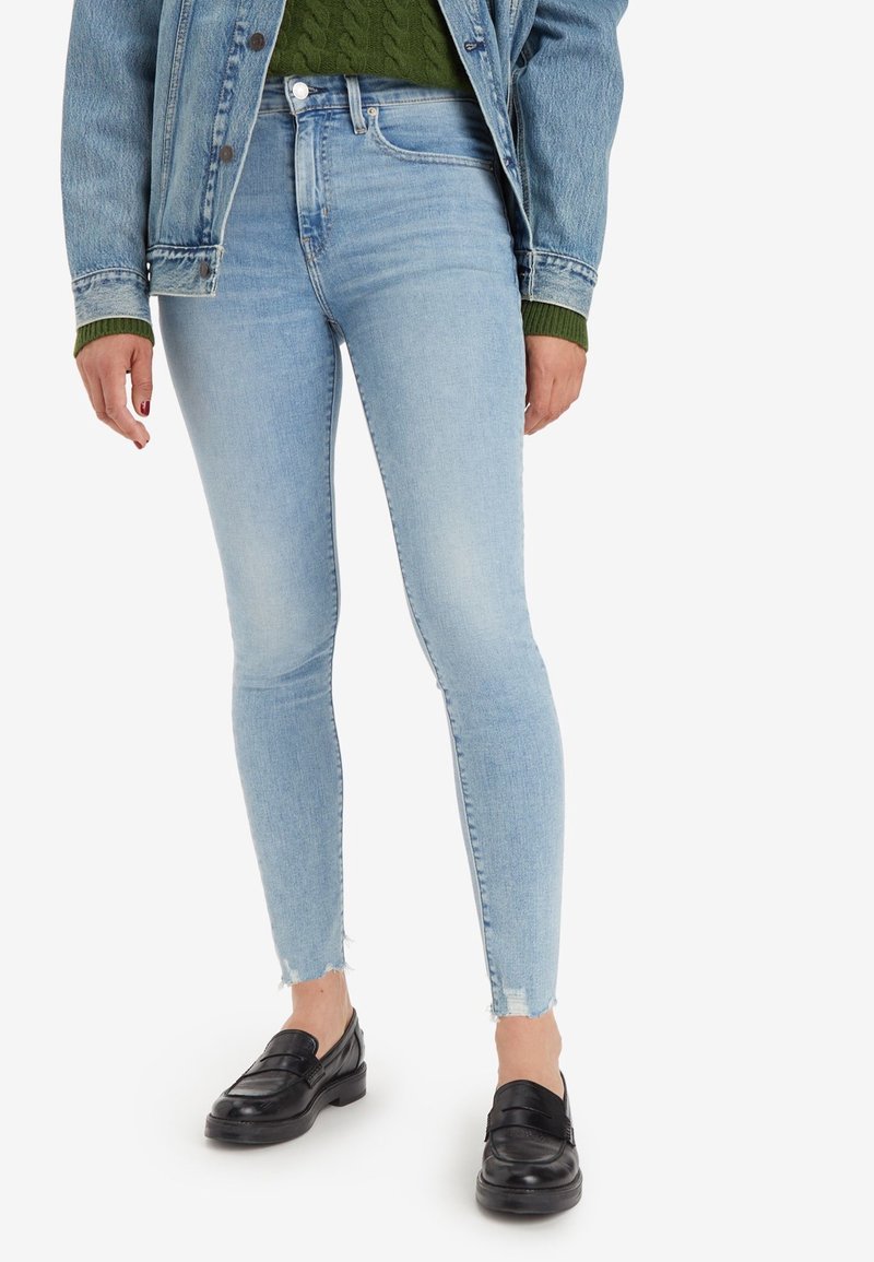 Femme portant un jean skinny bleu clair aux ourlets effilochés, des mocassins noirs, un pull vert et une veste en denim bleu, debout sur un fond blanc.