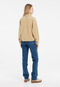 Beige fleecejakke med krave, knaplukkede ærmer og en afslappet pasform, parret med blå denimjeans og beige sko, set fra bagenden.