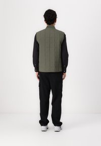 Grüner gesteppter Weste mit hohem Kragen, getragen über einem schwarzen Button-up-Hemd. Hat eine lockere Passform und wird mit schwarzen Cargo-Hosen und Sneakers kombiniert.