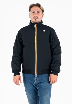 Giubbotto puffer nero con colletto alto, zip gialla a contrasto, polsini a costine e un piccolo logo sul petto, indossato sopra a jeans blu.