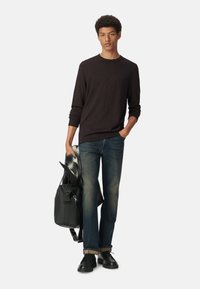 Schwarzes Langarmshirt, blaue Jeans mit fransigen Säumen, schwarze Schuhe und eine schwarze Umhängetasche, die einen grau-weißen Schal hält.