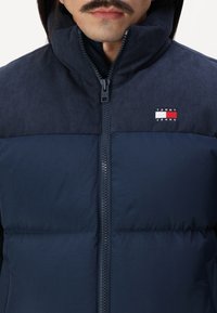 Marineblå pufferjakke med høy krage, horisontalt ribbet øverste del og glidelås. Har "TOMMY JEANS"-logo på brystet.