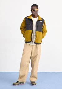 Giacca in pile con blocchi di colore giallo e grigio scuro, con tasche zip, indossata sopra una camicia bianca, abbinata a pantaloni cargo beige e sneakers con motivo.