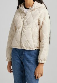 Světle béžová prošívaná bunda s kapucí, elastickými manžetami, předním zipem a bočními kapsami, vyznačující se hladkým, jemným povrchem.