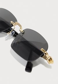 Occhiali da sole neri a forma quadrata con lenti scure, dettagli in hardware dorato e cuscinetti nasali trasparenti. Presentano una finitura liscia e un design geometrico.