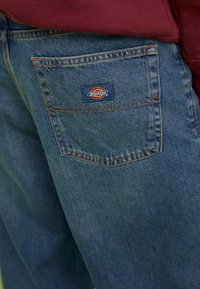 Blue denim jeans with prominent stitching, a back pocket featuring a red and yellow Dickies logo, and a relaxed fit.