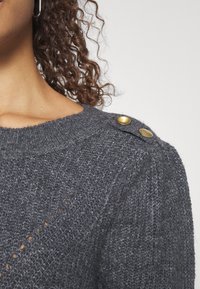 Pull en maille gris avec des accents de boutons dorés sur l'épaule, présentant un motif texturé et un large décolleté.