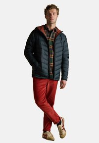 Mężczyzna w czarnej pikowanej kurtce, założonej na zieloną i pomarańczową koszulę w kratę, w czerwonych spodni od dżinsu i beżowych sneakersach, stojący z założonymi nogami.