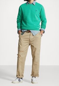 Grön sweatshirt, ljus denimskjorta, beige slitna byxor med hål och vita sneakers med svarta ränder. Avslappt lager-på-lager-outfit.