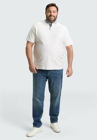 Weißes Poloshirt mit kurzen Ärmeln und blauem Kragen, kombiniert mit hellblauen Jeans und weißen Sneakers. Das Shirt hat eine geknöpfte Knopfleiste.