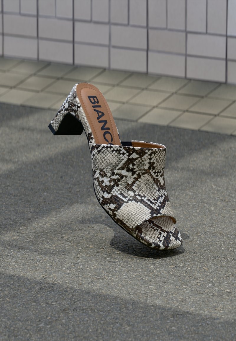 Ανοιχτό-toe mule με block τακούνι και μοτίβο φίδιου δέρματος, με ορατό όνομα μάρκας στην εσωτερική σόλα, τοποθετημένο σε υφή εξωτερικής επιφάνειας.