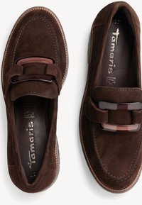 Braune Wildlederslipper mit runder Zehenform, ausgestattet mit einem doppelten Riemenakzent in glänzender Optik. Lederinnensole mit Logo-Detail und Steppnaht.