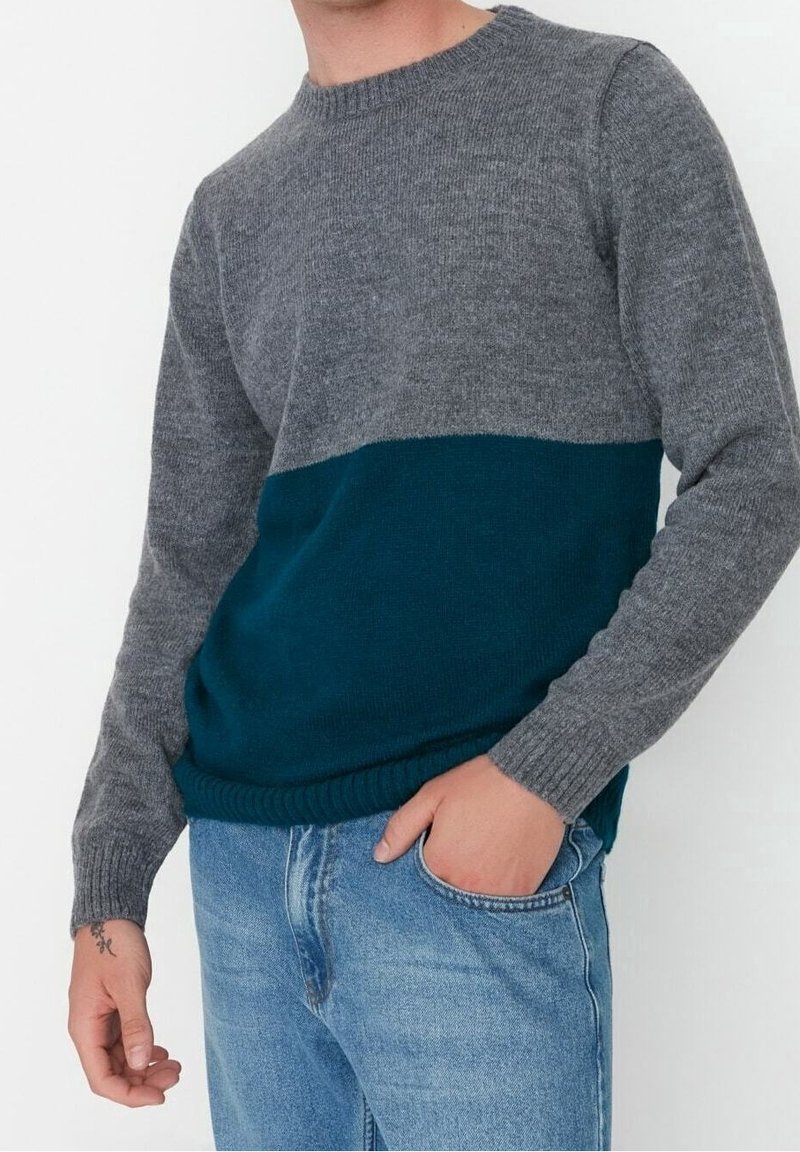 Gebreide trui met een grijs bovenstuk en een teal onderstuk, ronde halslijn, geribbelde mouwen en zoom, en een zachte textuur. Gecombineerd met lichtblauwe jeans.