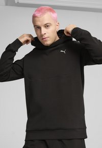 Sort hættetrøje lavet af glat stof; har en frontlomme og et lille hvidt Puma-logo på brystet. Modellen poserer med hænderne ved hætten.