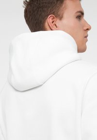 Felpa bianca con cappuccio in tessuto morbido e testurizzato; presenta un cappuccio arrotondato e un design semplice, vista dal retro.