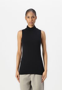 Haut sans manches en turtleneck noir en tissu doux et extensible ; design ajusté avec un col haut, associé à un pantalon beige clair.