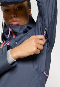 Ναυτικό σακάκι με ψηλό γιακά, τσέπες με φερμουάρ και αντίθετα φερμουάρ σε πορτοκαλί και μωβ χρώμα. Διαθέτει υφή υφάσματος και ρυθμιζόμενα στοιχεία.