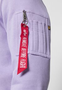 Lilla sweatshirt med en lynlås lomme, tekstureret lap og et rødt "REMOVE BEFORE FLIGHT"-mærke fastgjort til lynlåsen.