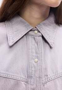 Κοντινό πλάνο ενός ατόμου που φορά ένα ανοιχτό μωβ τζιν μπουφάν με κουμπιά πέρλες και εμφανείς ραφές στον γιακά και στο στήθος.