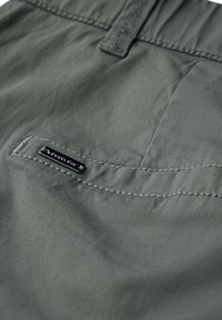 Ciemnozielony materiał tekstylny z widocznymi szwami, z tylną kieszenią i małą czarną metką logo przymocowaną do tkaniny.