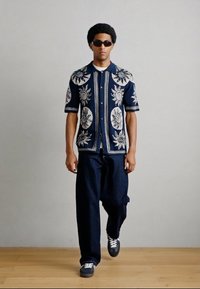 Μπλε σκούρο πουκάμισο με κοντά μανίκια και περίπλοκη λευκή κεντήματα λουλουδιών, συνδυασμένο με άνετο σκούρο τζιν παντελόνι και μαύρα αθλητικά παπούτσια.