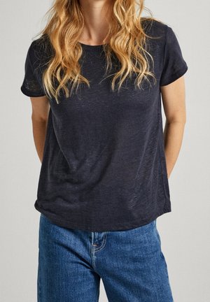 Personne blonde portant un t-shirt noir uni à manches courtes, non rentré, sur un jean bleu, sur un fond neutre.