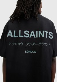 Juodas medvilnės marškinėlis su dideliu baltu tekstu ant nugaros: "ALLSAINTS", su papildomu tekstu japonų kalba ir "LONDON" apačioje.