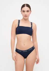 Navy bikini komplet s bandeau topom s dvostrukim trakama i Calvin Klein logom, upotpunjen odgovarajućim hipster donjim dijelom s elastičnom trakom s logom.