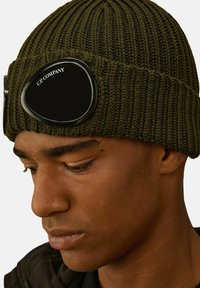 Beanie a coste color verde oliva con dettagli neri a forma di occhiali, caratterizzato da una texture liscia, una forma distintiva e un logo sui goggles.