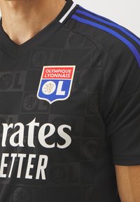 Maillot de football noir de l'Olympique Lyonnais avec un blason rouge, blanc et bleu et des bandes bleues sur l'épaule, porté par une personne.