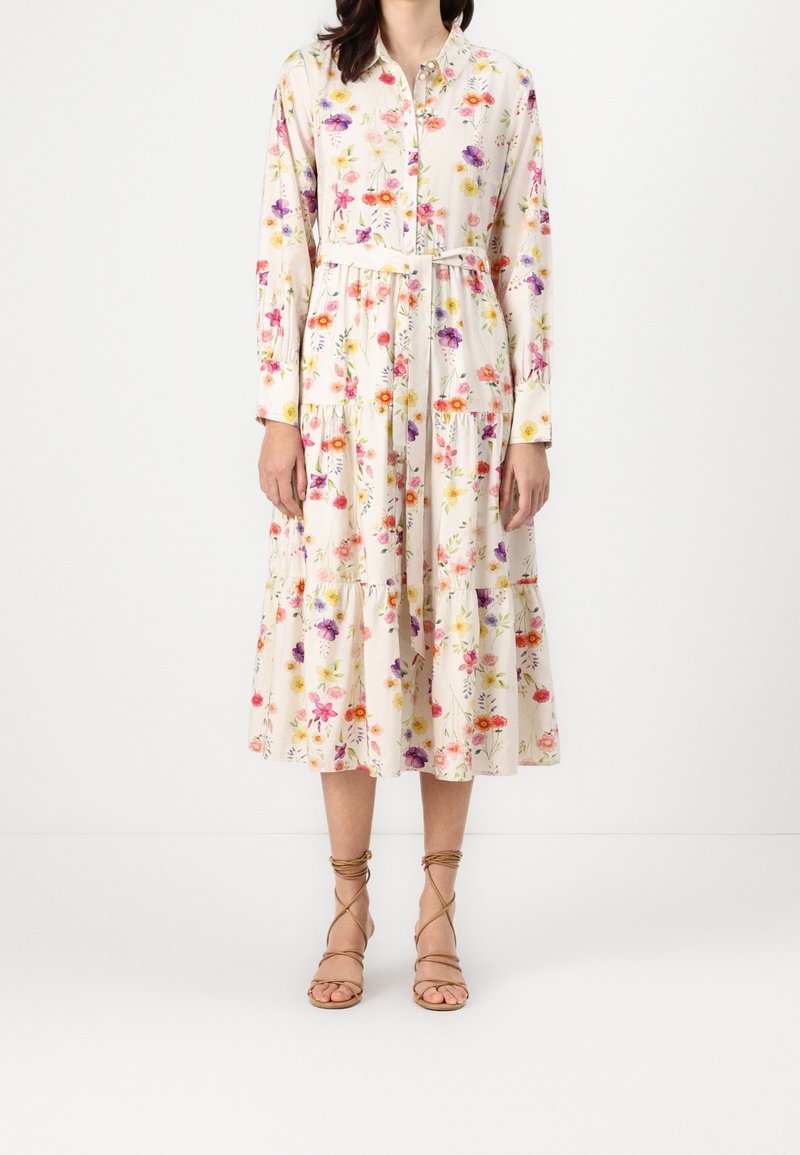 Femme portant une robe midi crème avec un imprimé floral coloré, des manches longues, des boutons sur le devant, une ceinture à la taille et des sandales à lacets couleur beige.