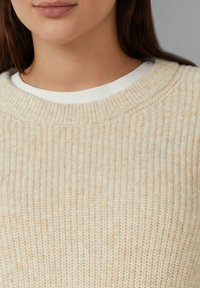 Beżowy sweter dziany z ryżowym wzorem i okrągłym dekoltem, noszony na białej koszulce z okrągłym dekoltem.