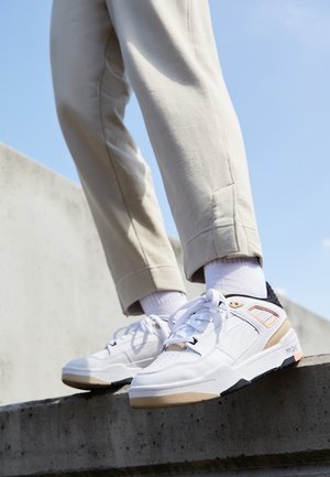 Des chaussures de sport blanches avec une surface texturée, accentuées de crème et de noir. Elles présentent une semelle proéminente et un design lacé classique.