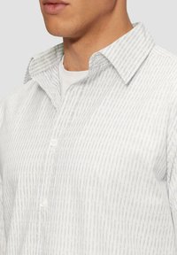 Witte blouse met een subtiele paarse streepprint, voorzien van een gespreide kraag en een textuurstof, voor een deel zichtbaar op een model.