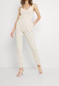Beige jumpsuit met een textuurrijke, gebreide buste. Elastische tailleband, zijzakken en taps toelopende benen. Gepaard met witte sandalen met bandjes.