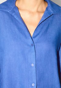 Blaues, kragenloses Hemd aus leichtem Stoff, das eine glatte Textur aufweist und passende Knöpfe auf der Vorderseite hat.