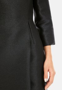 Schwarzes Kleid mit subtiler Schimmer, strukturiertem Stoff, taillierter Passform und einer seitlichen Falte. Eine Hand mit natürlichem Nagel Finish liegt auf dem Stoff.