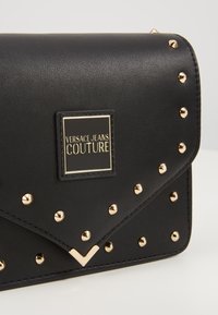 Borsa a tracolla in pelle nera con borchie dorate, una placca con logo al centro e un design distintivo con pattina che presenta un accento a forma di V.