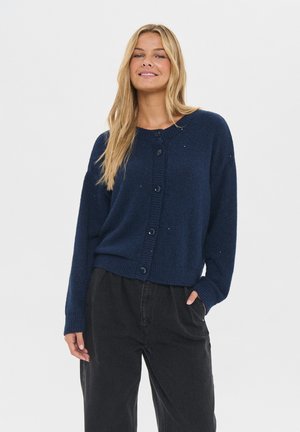 Lächelnde Frau mit langen blonden Haaren, die einen navy-blauen, knopfgeschlossenen Pullover und lockere schwarze Hosen trägt, während sie mit einer Hand in der Tasche posiert.