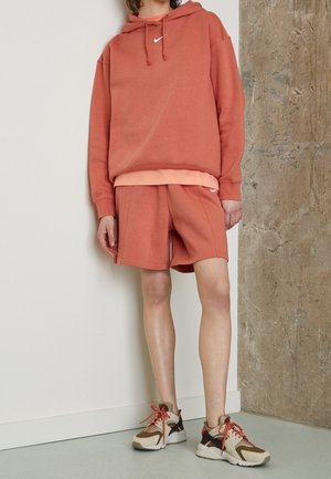 Personne portant un sweat à capuche et un short assortis couleur rouille avec des baskets beige et marron, debout contre un mur à texture béton.