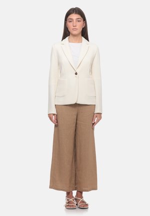 Donna in piedi che indossa un blazer bianco testurizzato sopra una canotta bianca, pantaloni marroni a gamba larga e sandali bianchi con cinturini, rivolta in avanti su sfondo bianco.