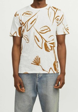 Homme portant un t-shirt blanc avec un grand imprimé feuille marron et fleur, associé à un jean en denim bleu clair.