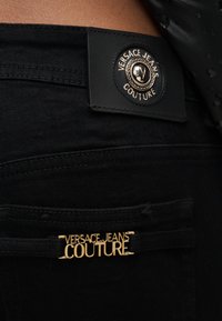 Κοντινό πλάνο μαύρου παντελονιού Versace Jeans Couture που δείχνει την χρυσή πλακέτα με το λογότυπο στην πίσω τσέπη και μαρκαρισμένη ετικέτα πάνω από τη μέση.