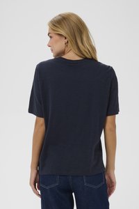 Tamsiai mėlynas trumpomis rankovėmis marškinėliai su apvaliu iškirpimu, laisva siluetu ir minkšta tekstūra. Šiek tiek ilgesnis nugaros hemelis su sukamu rankoviu dekoracijomis.