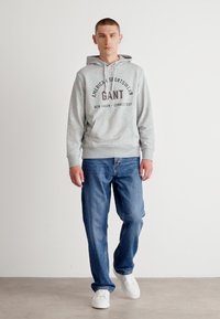 Sudadera gris con el texto "GANT", combinada con jeans azules y zapatillas blancas. Conjunto casual de corte relajado y diseño estándar de sudadera con capucha.